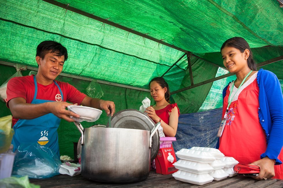 Maly buys lunch from CARE’s food vendor in front of her factory. “Wejoined CARE’s program to learn how to improve our cooking and hygiene”, says Veasna, who has been cooking and selling his food in front of the factories for months. Phnom Penh, Cambodia. Mar. 14, 2018. © CARE / Erika Piñeros
