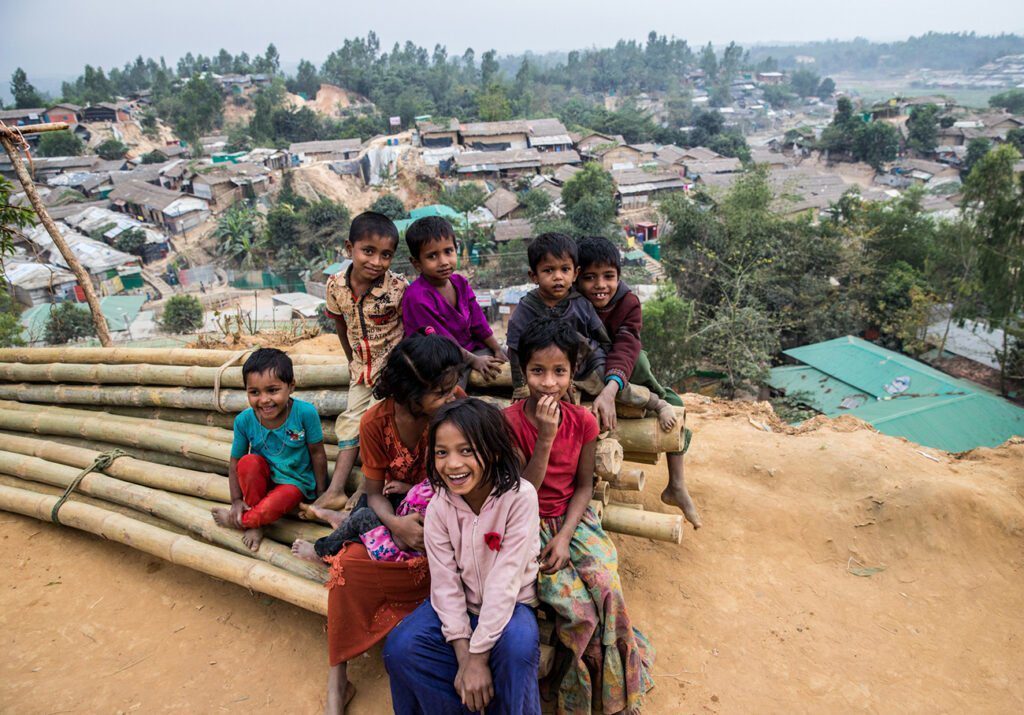 A group of children smile from a bamboo pile meant for home repairs, overlooking the Palangkhali refugee camp in Bangladesh..