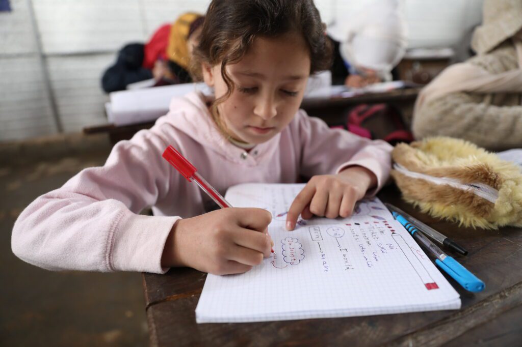 A young girl, Hana, sits at a desk writing in a notebook. Shafak/CARE
