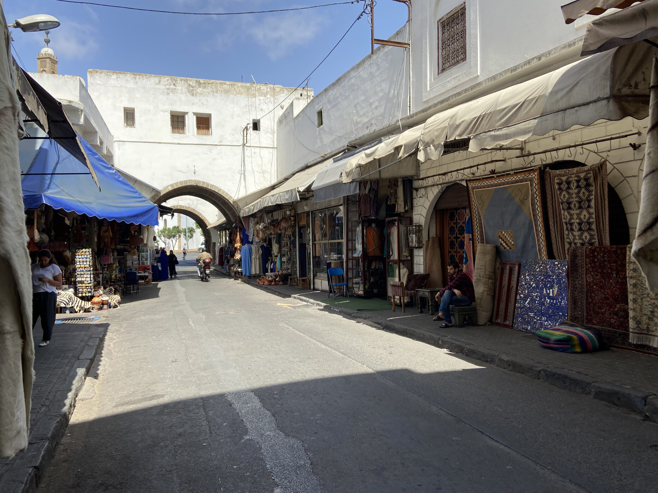 Photo taken from the middle of a street. Shops down the right hand side with various textiles.