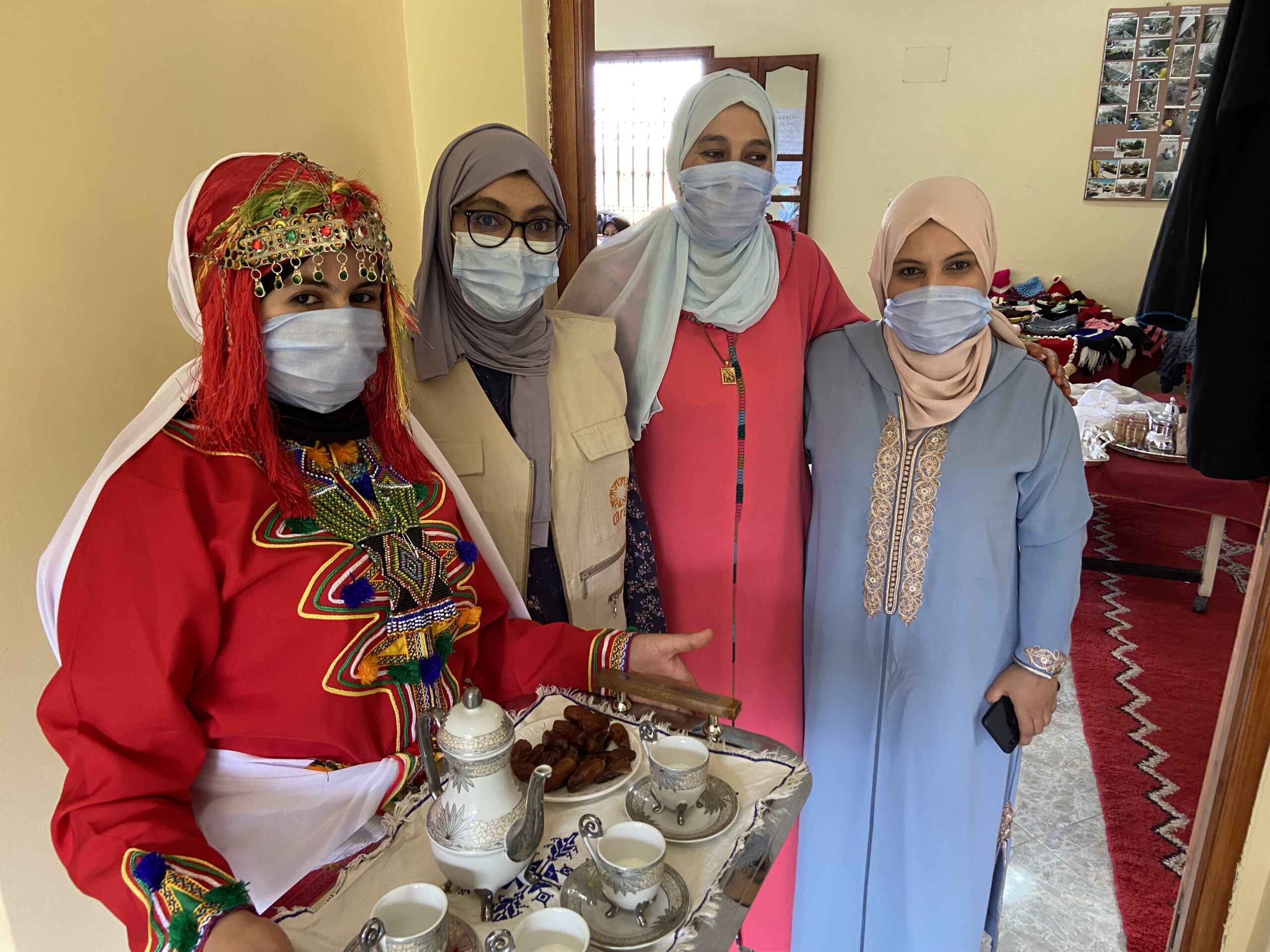 4 women standing together in a line for a photo. The woman on the far right is holding a tea tray with a tea pot and various items on it.