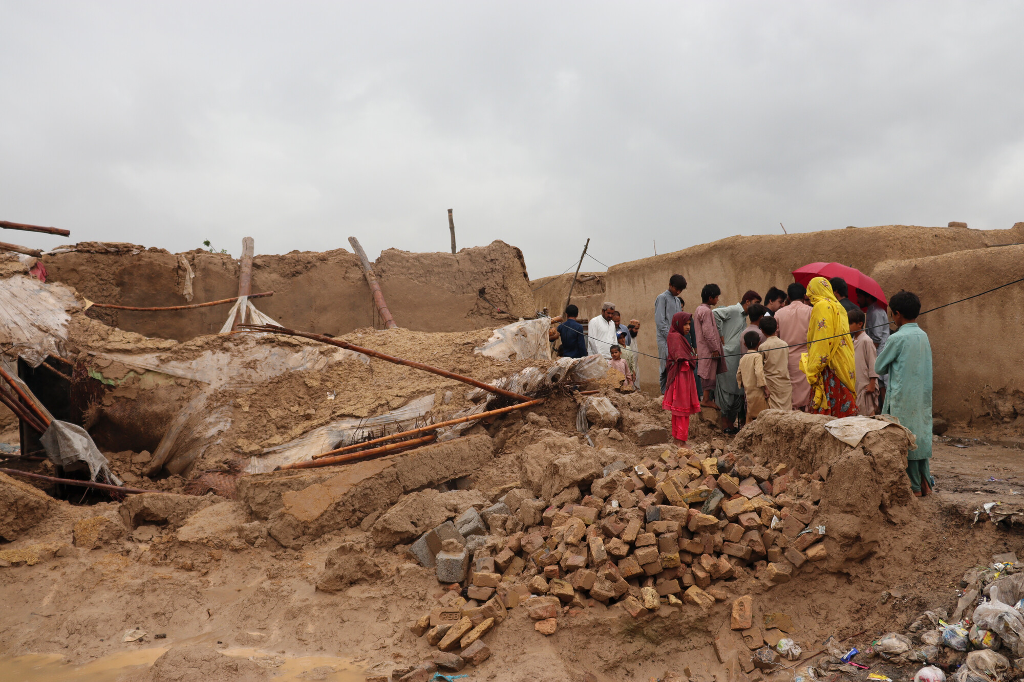 A group of people stand together near a pile of rubble and the remains of a home after floods destroyed it.