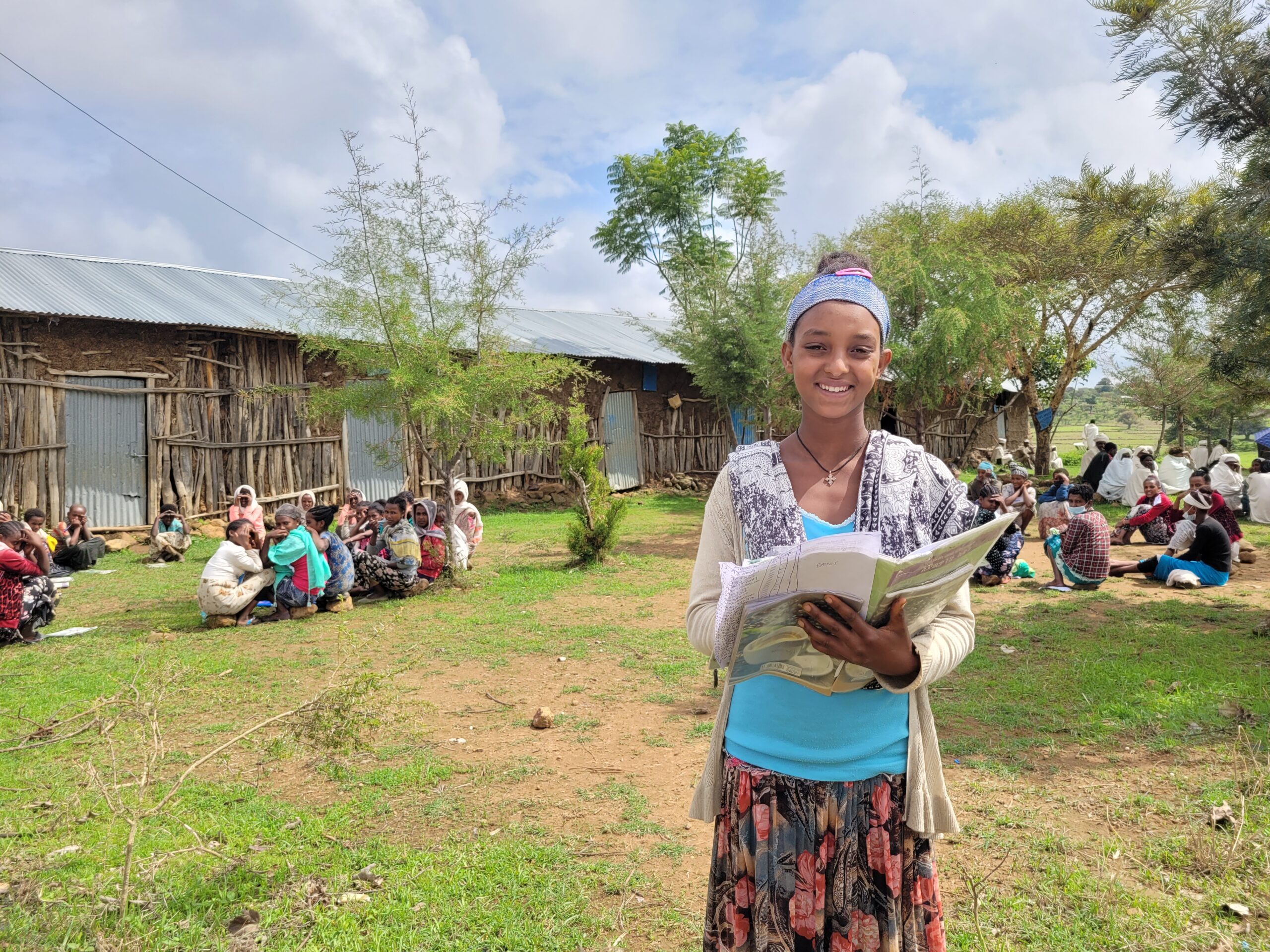 A girls smiles brightly while she stands outdoors holding an open book. Behind her a group of girls site in a circle.