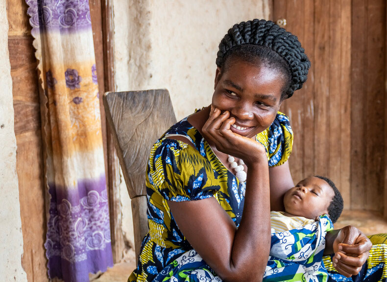 A woman sits on a chair leaning her chin on her palm as she smiles warmly. She holds a small baby in her other arm.