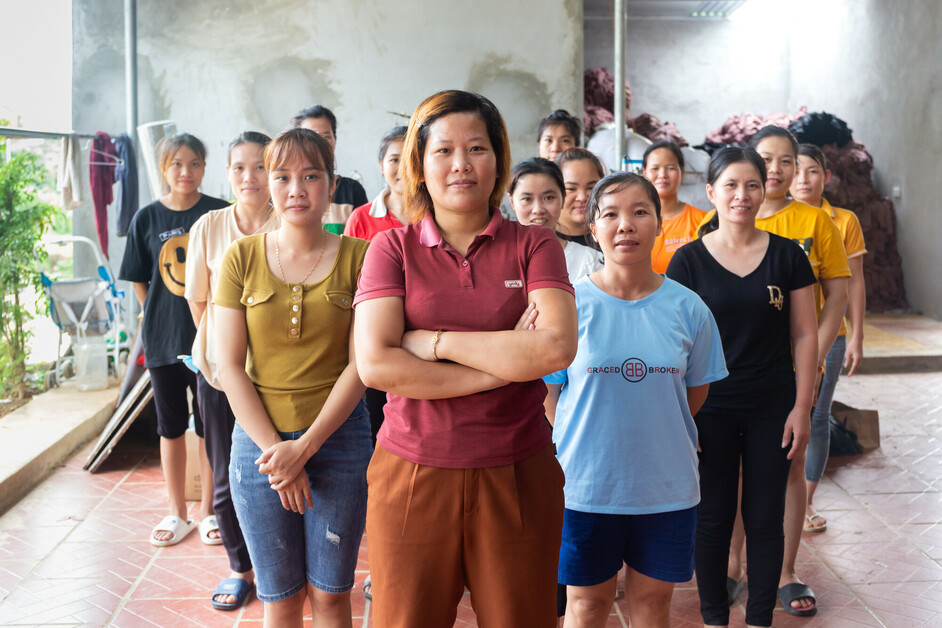 A group of women stand together facing forward in a room. The woman in front stands with her arms crossed.