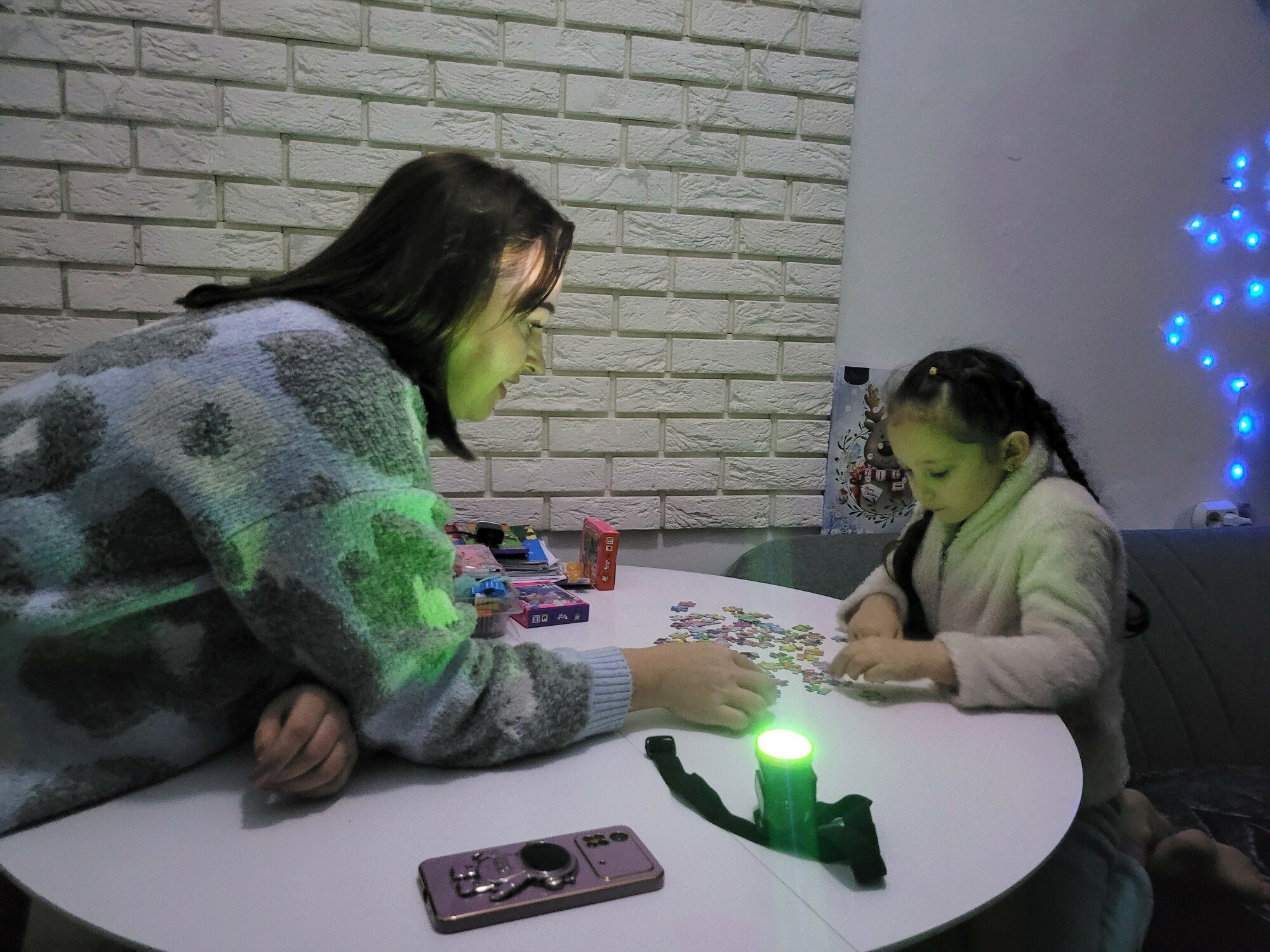 A other and daughter sit at a table doing a puzzle using a flashlight to see
