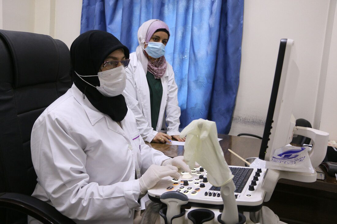 A woman wearing a mask and a white coat sits at an ultrasound machine. Another woman in a white coat stands beside her