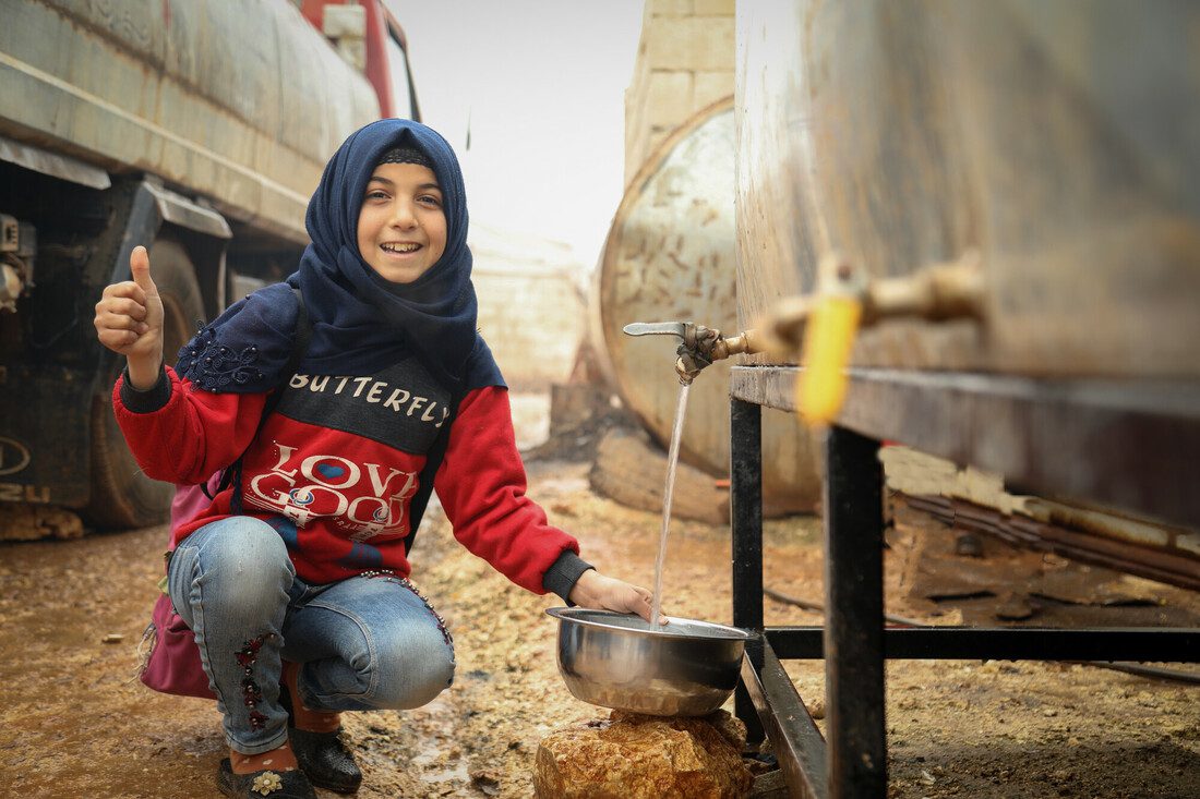A girl squats down to gather water from a tank with a tap. She smiles and holds her thumb up. Behind her are other tanks in a refugee camp.