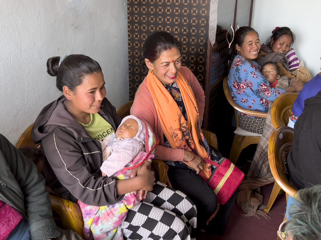 Four women sit on chairs in a building. Two of the women are holding babies and smiling.