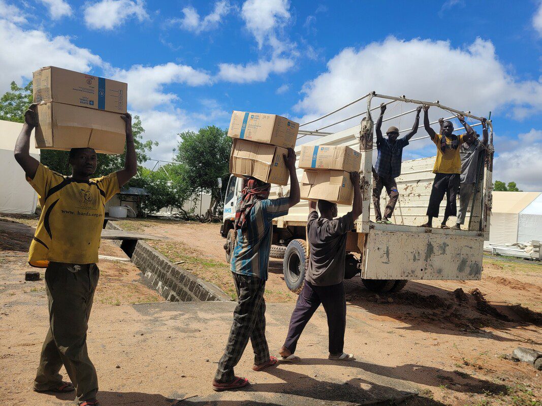 Three men walk outdoors carrying boxes on their heads towards a truck where people are standing.