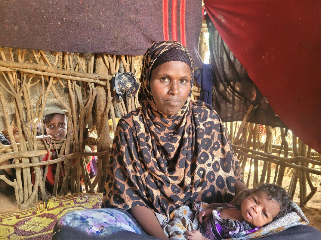 A woman sits in a straw hut while holding a baby. A young child stands behind her