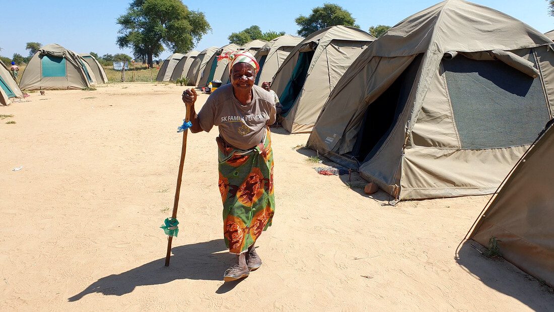 An elderly woman walks with a stick. The backdrop behind them is sunny, dry and sandy and there are other tents around.