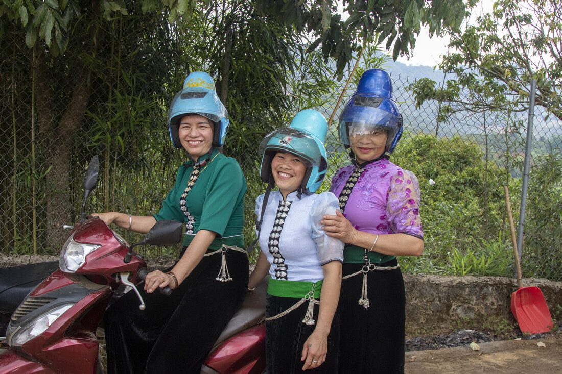 3 women stand beside a red scooter. They are all wearing the special helmets that allow for their 6 inch buns.