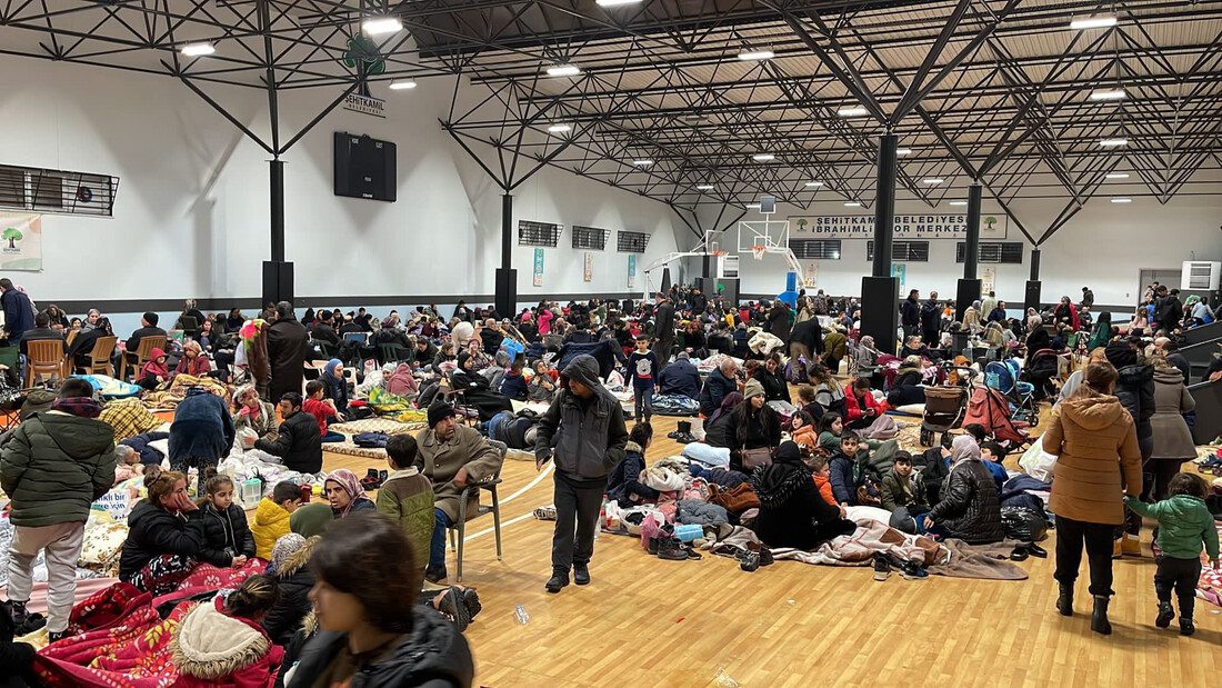 A large room indoors crowded with people and luggage, bedding, tables and chairs.