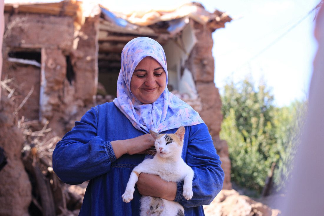 A woman holds a cat and smiles down at it. She stands in front of a damaged home.