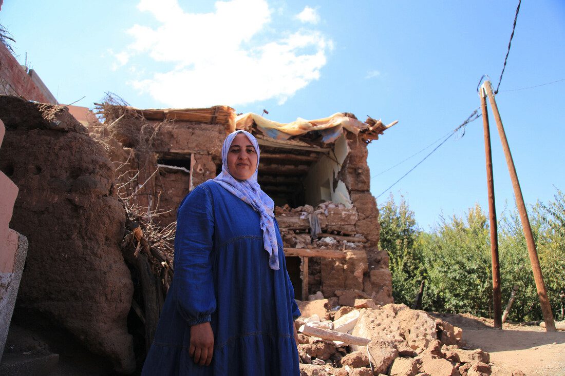 A woman stands outdoors. Behind her is a damaged home and rubble on the ground.