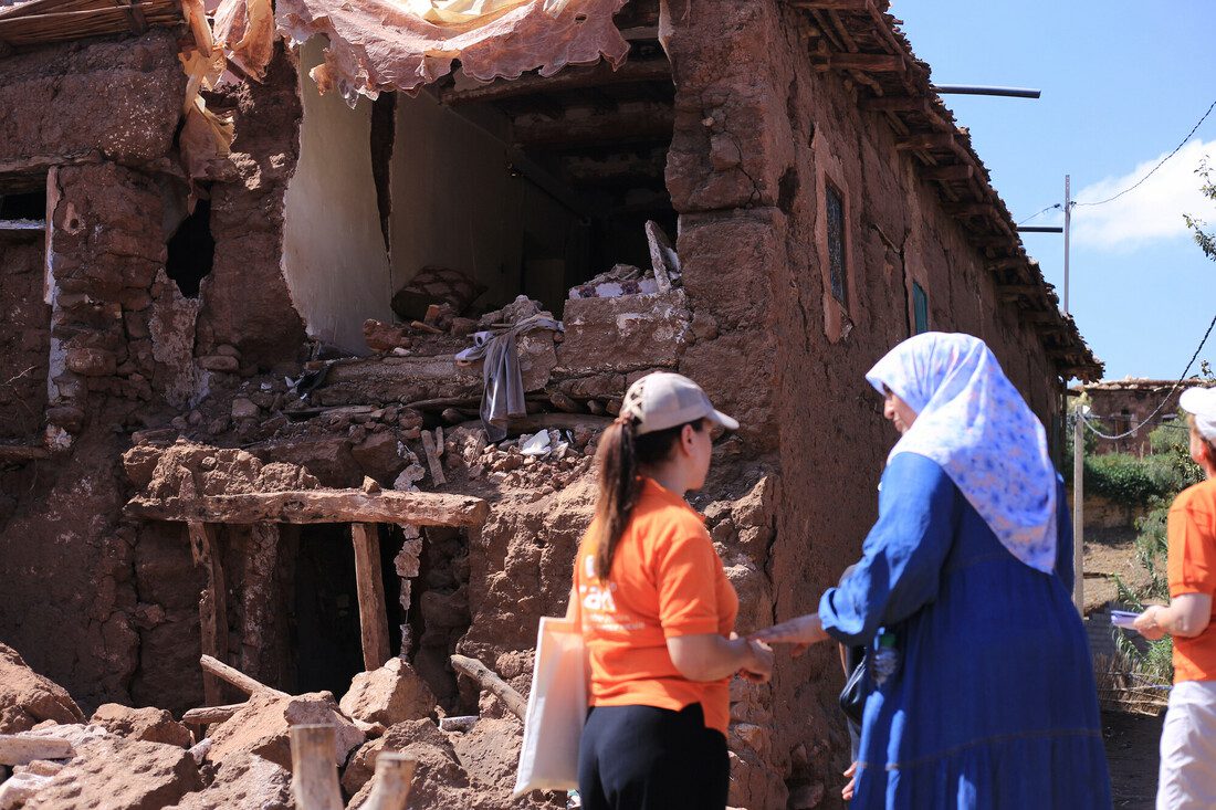 Two women stand together outside. One is wearing a CARE-banded shirt. The building behind them is badly damaged from an earthquake. Debris and rubble lie around.