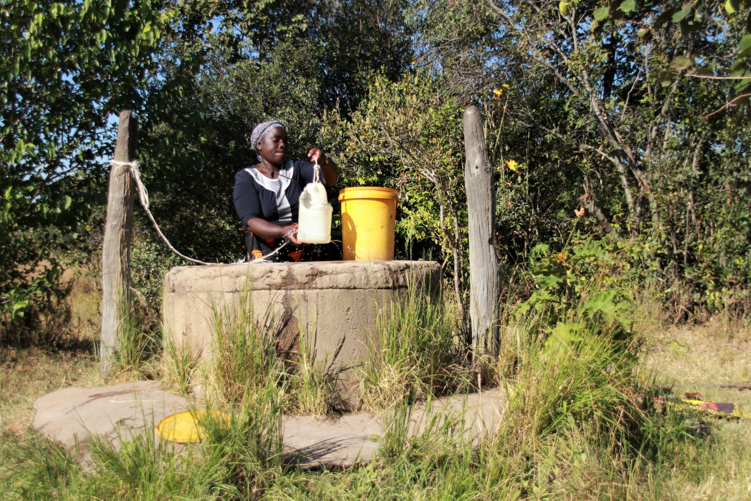 A woman stands outside behind a large stone well. She uses a small white container, attached to a rope, to collect the water and fill a larger yellow bucket beside her.