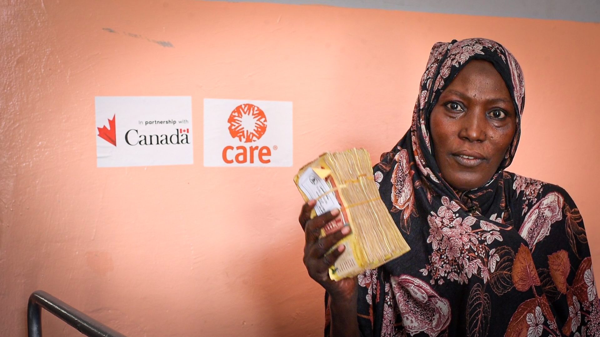 Une femme tient une liasse d’argent dans une main. Elle est devant un mur orange affichant le logo de CARE et celui du gouvernement du Canada.