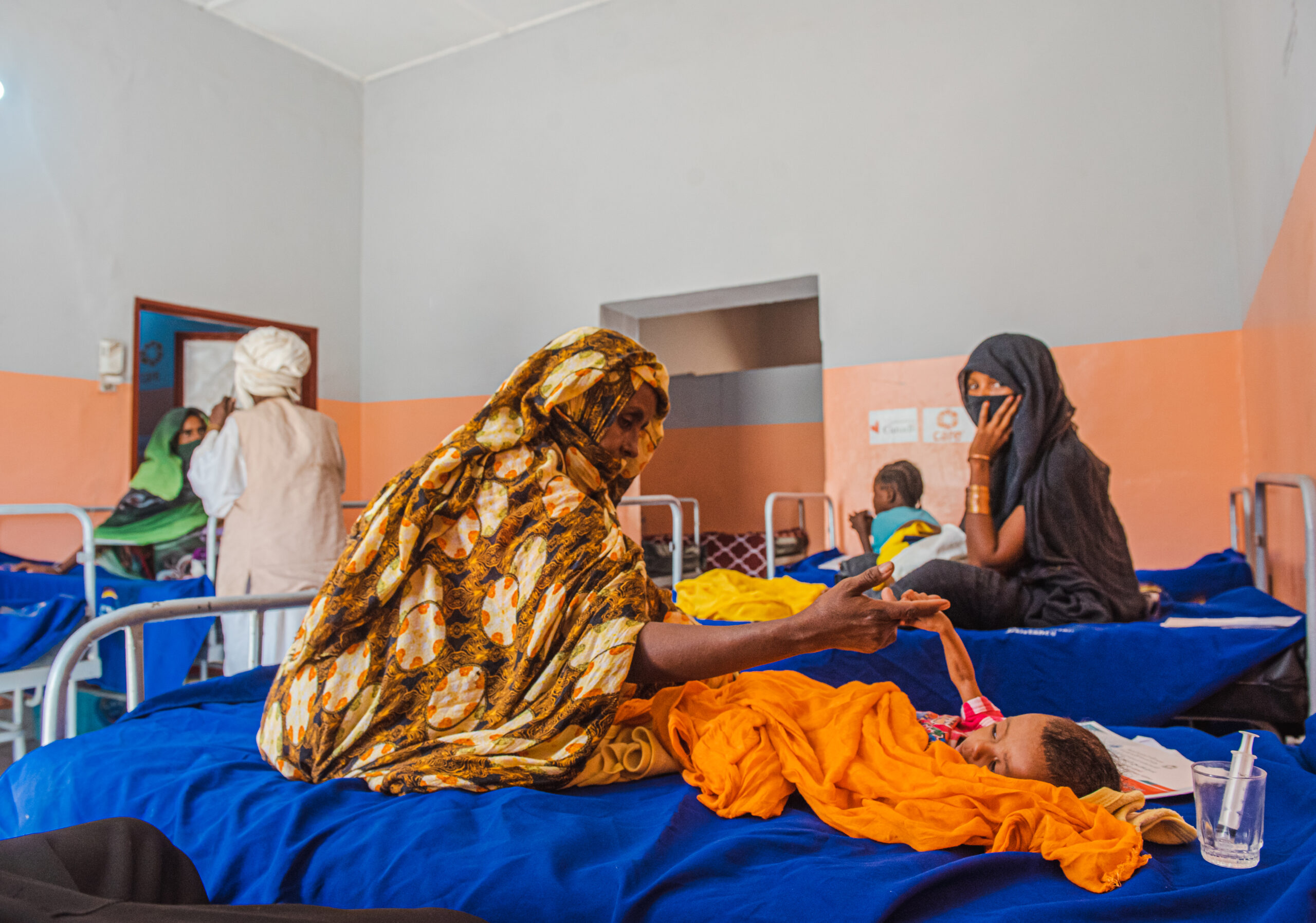 A woman sits on a hospital bed, holding the hand of a young child who is lying down.