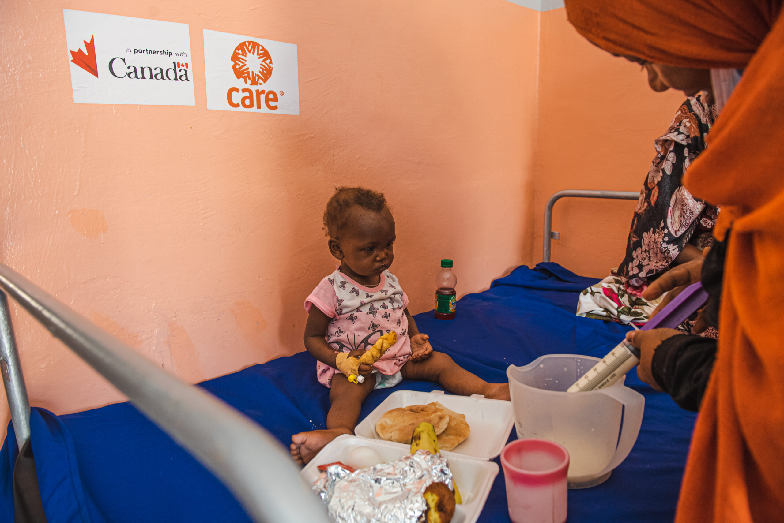 An infant sits on a hospital bed with food in front of her. She watches a woman who is standing beside the bed fill a large syringe with white liquid from a plastic container.