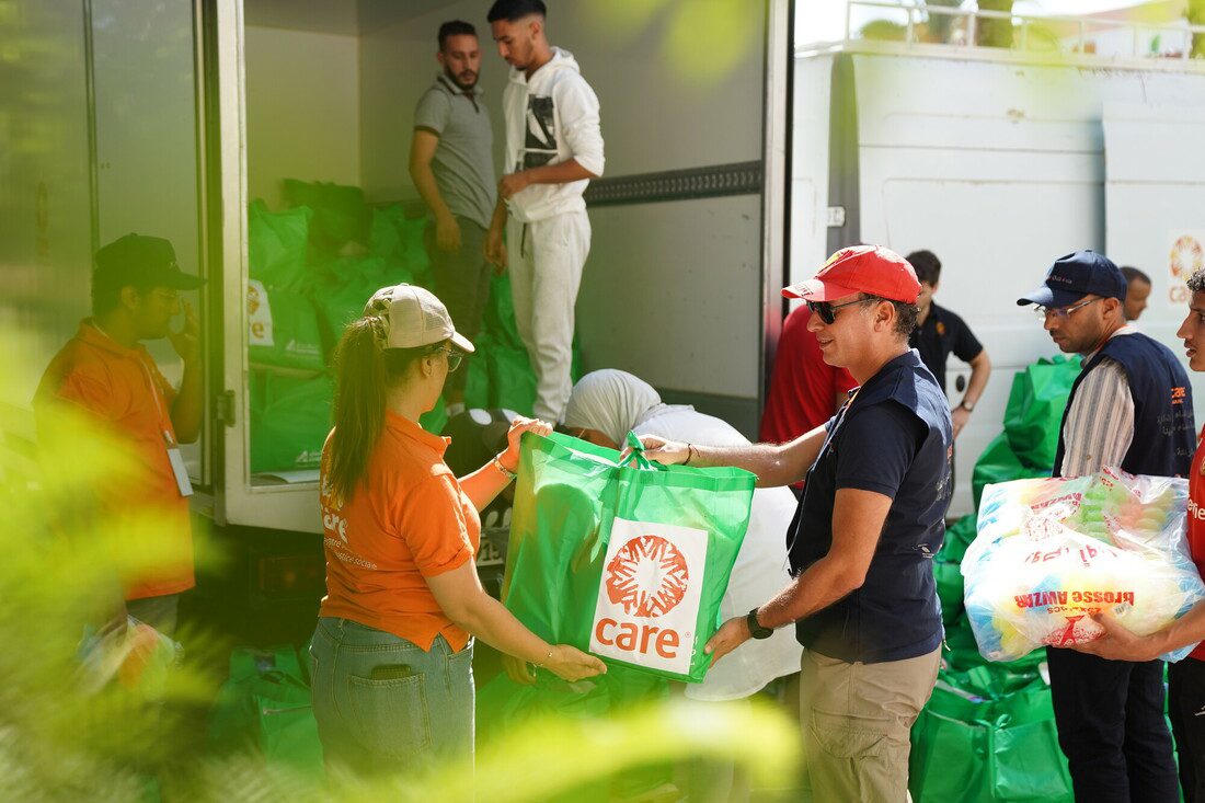 A woman wearing an orange CARE shirt passes a green CARE package to a man. They are standing in front of an open truck that is full of CARE packages. 