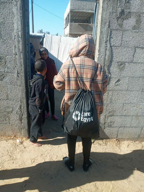 A person faces a grey stone wall. They wear a coat with a hood pulled up and a black draw string CARE bag on their back. 