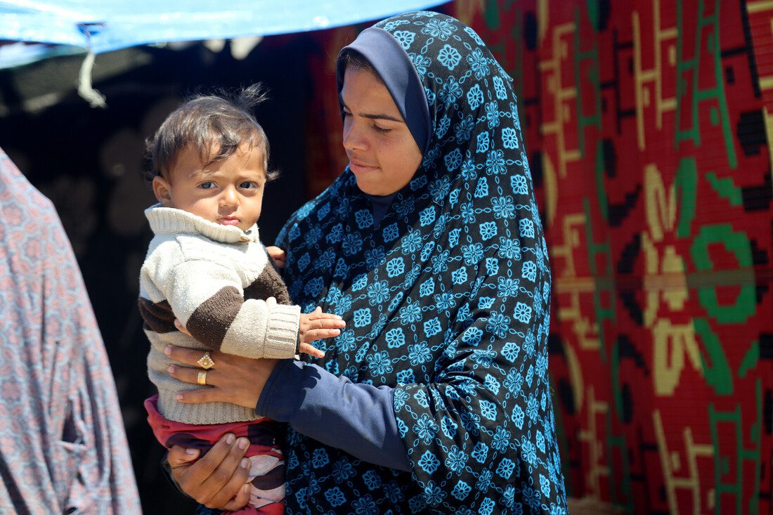 A woman wearing a blue hijab stands holding a young child in her arms.