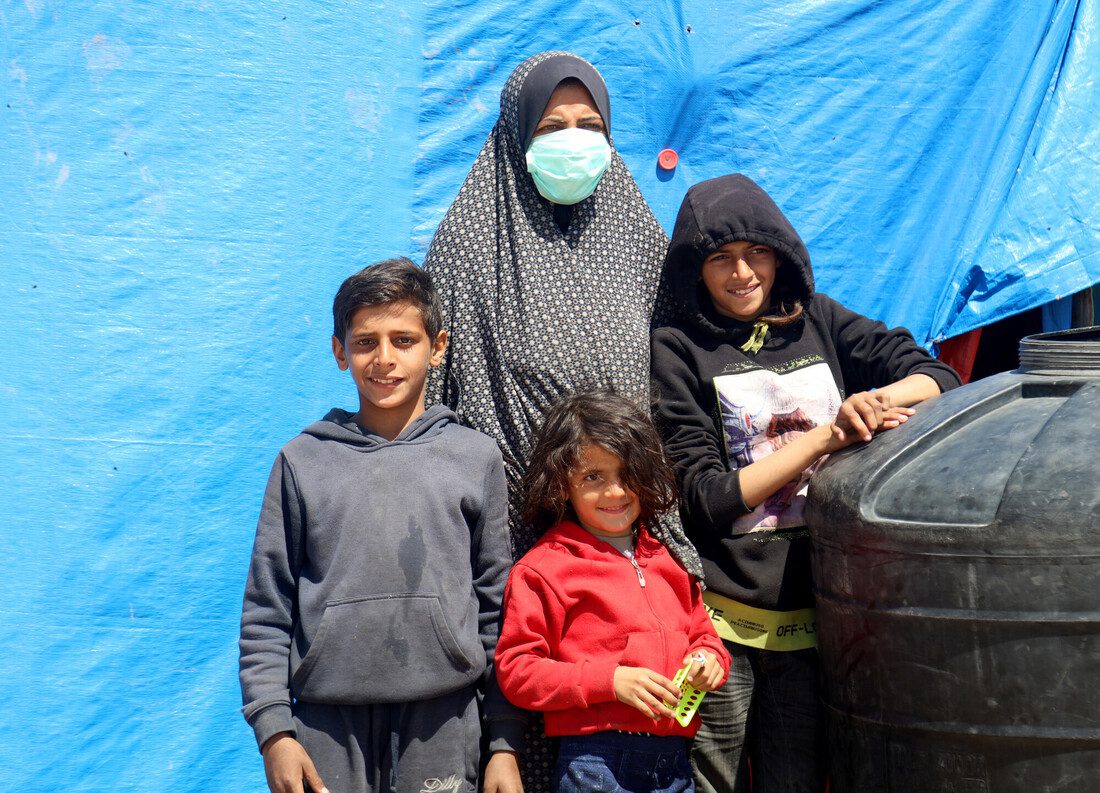 A woman stands with her three children in front of a blue tarp. Beside them is a large black container 