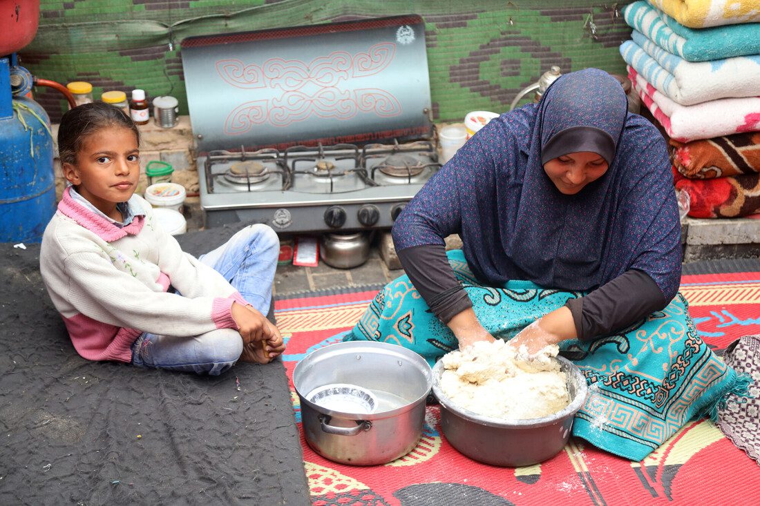 A woman sits on a colourful rug, preparing food. A young girl sits beside her on a cot. 