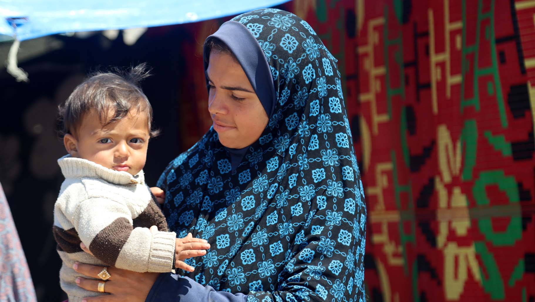 A woman wearing a blue hijab stands holding a young child in her arms.