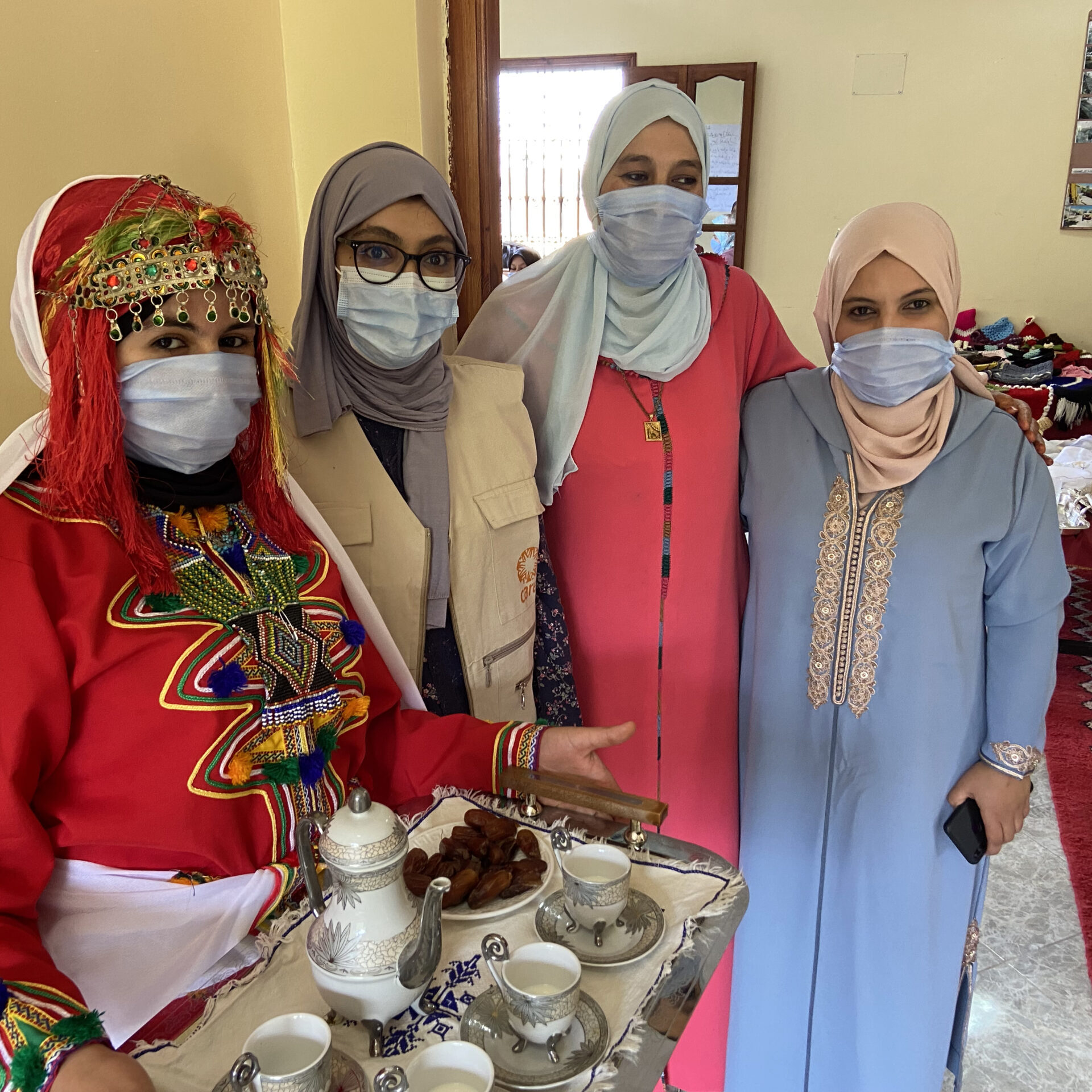 4 women standing together in a line for a photo. The woman on the far right is holding a tea tray with a tea pot and various items on it.
