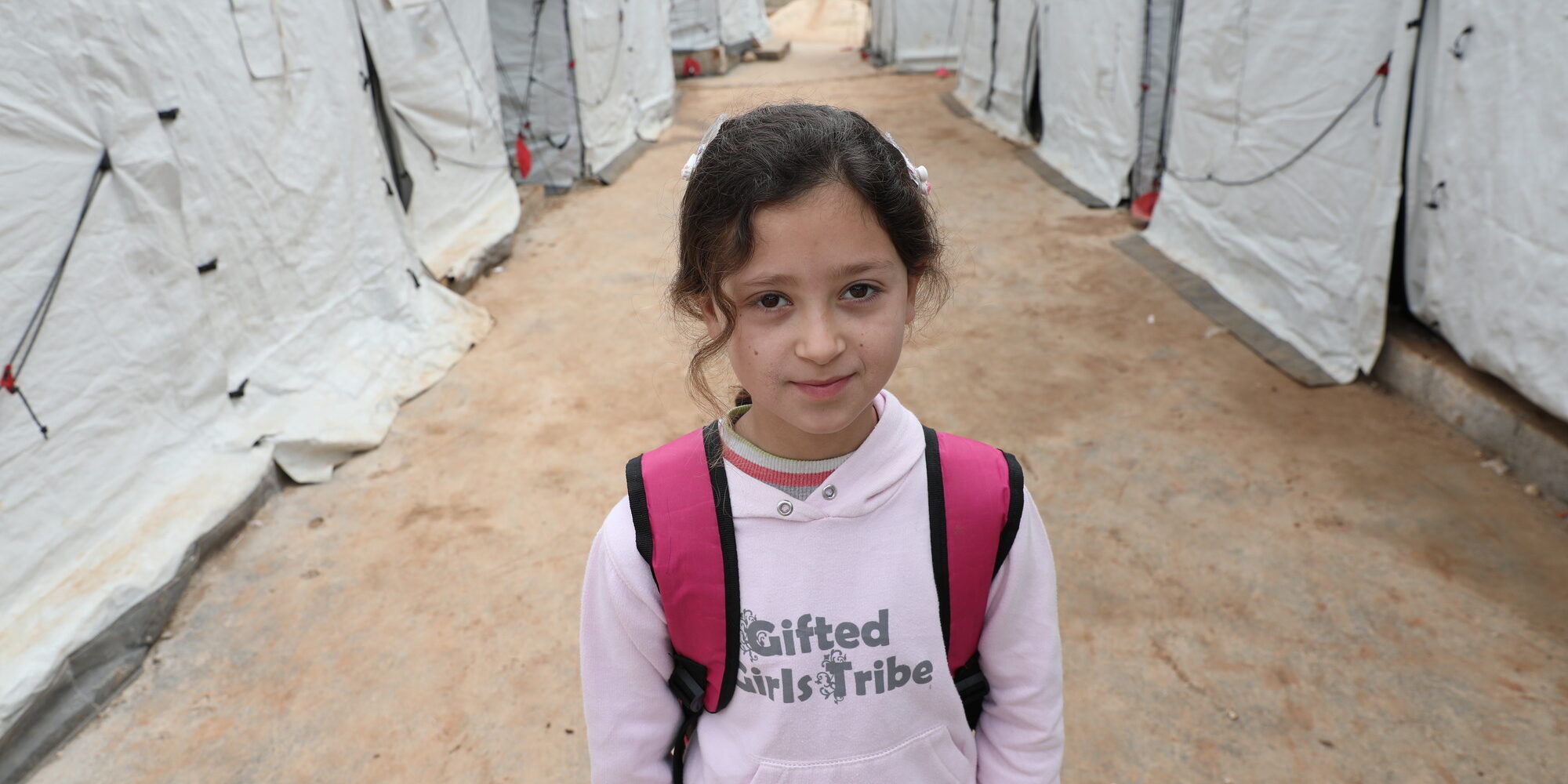A young girl, Hana, stands amidst a row of tents wearing her backpack in Azraq refugee camp. Shafak/CARE